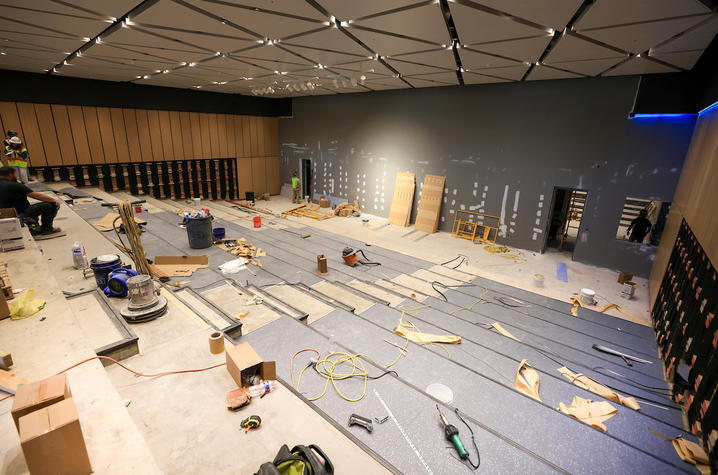 An auditorium under construction with tiered seating, scattered tools and materials, and workers installing fixtures.
