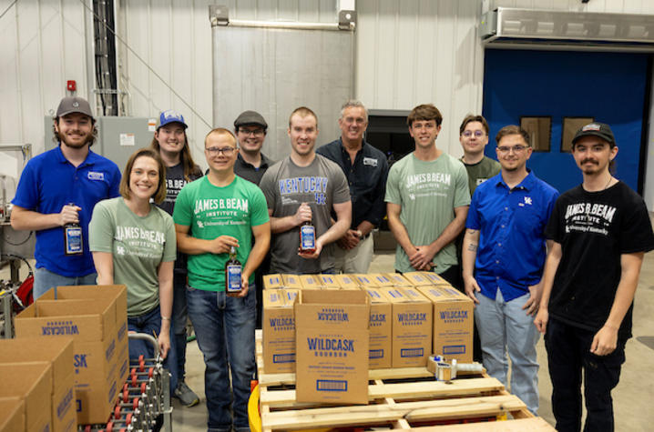 Wildcask Bourbon student creators pose in the bottling facility behind a pallet containing cardboard boxes with the brand name on them