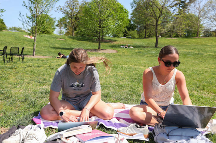 Two students sit on a blanket in a grassy park on a sunny day, studying and working on laptops with notebooks and supplies spread around them, while other people relax in the background.