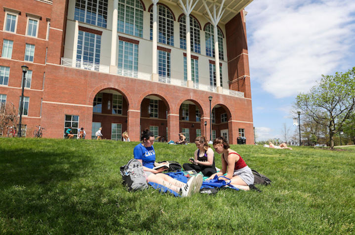 Three college students sit on a grassy lawn studying together outside a large brick academic building with tall arched windows, while other students relax in the background on a sunny day.