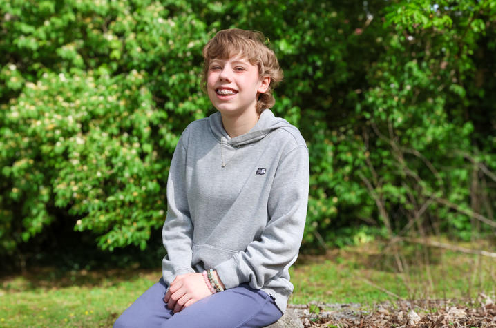 A young person sits on a low stone or log outdoors, wearing a gray hoodie and dark pants, with leafy green trees filling the background.
