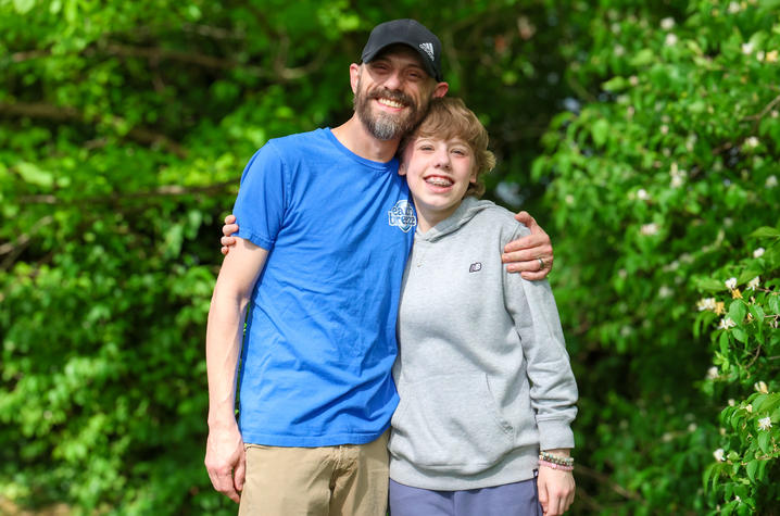 Two people stand outdoors in front of green trees, with one person’s arm around the other’s shoulders. One wears a blue T-shirt and cap, the other a gray hoodie.