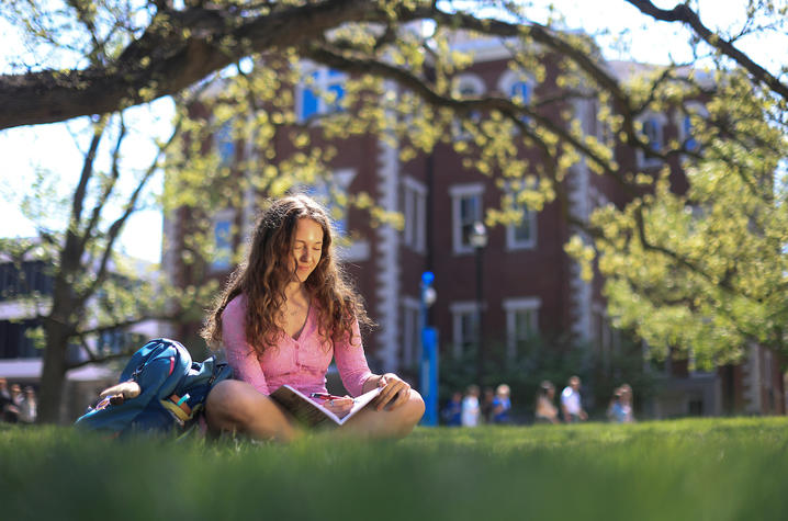Person sitting on a grassy lawn in front of a brick campus building, reading an open book with a backpack nearby and trees framing the scene.
