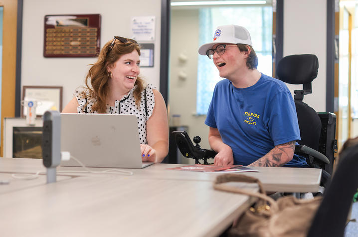 Two people seated at a table during a meeting, one using a wheelchair, with a laptop and other equipment on the table inside an office or classroom setting.