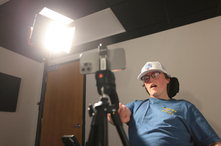 Person seated in a wheelchair looks toward a camera on a tripod under bright studio lights in an indoor recording space.