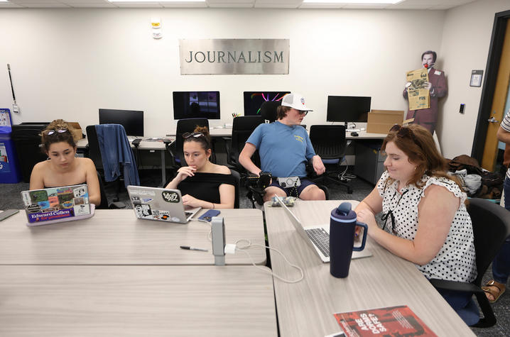 Five people sit around a conference table working on laptops in a classroom labeled “Journalism,” with desktop computers along the walls.