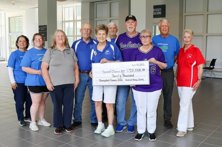 A group of adults stands indoors in a lobby, posing together while two people hold a large ceremonial check for $20,000 made out to Second Chance KY. 