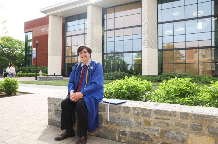 A graduate wearing a blue cap and gown sits on a stone bench outside an academic building with large glass windows and brick accents. Green shrubs and a campus walkway surround the seating area in daylight.