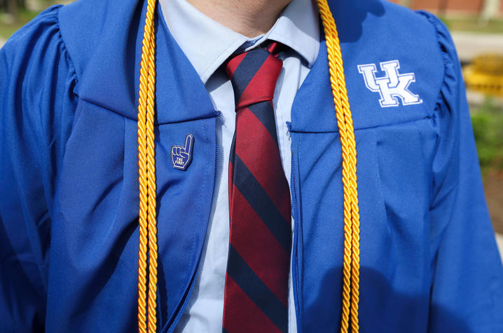Close‑up view of a University of Kentucky blue graduation gown worn over a dress shirt and red‑and‑blue striped tie, with gold honor cords draped around the neck and a small lapel pin visible on the gown.