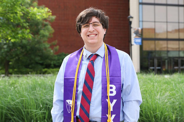 A graduate stands outdoors on campus wearing a blue graduation gown over a dress shirt and red‑and‑blue striped tie, with a purple and gold academic stole draped around the neck. 