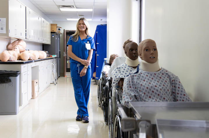 A person wearing blue medical scrubs stands in a clinical skills hallway lined with wheelchairs holding medical training manikins dressed in hospital gowns, with storage cabinets and additional manikins visible in the background.