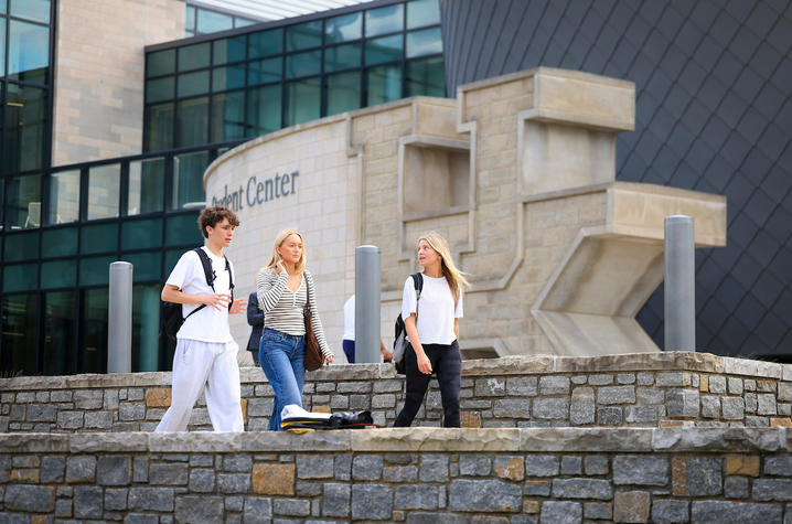 People walking and talking on a sidewalk near a large stone “UK” sculpture outside a modern campus building.
