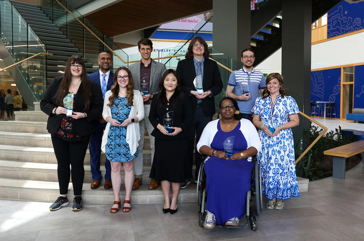 A group of University of Kentucky faculty and graduate teaching assistants stand and sit on indoor steps holding glass awards inside a modern campus building.