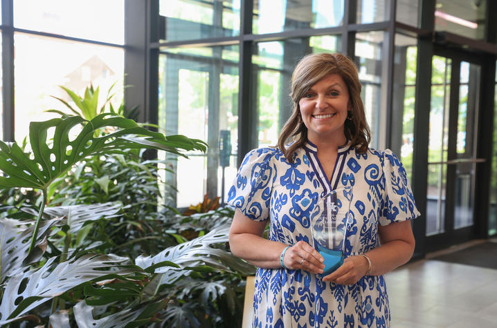 A person stands in a bright campus building lobby near large green plants, wearing a blue-and-white patterned dress, with glass windows and doors visible in the background.