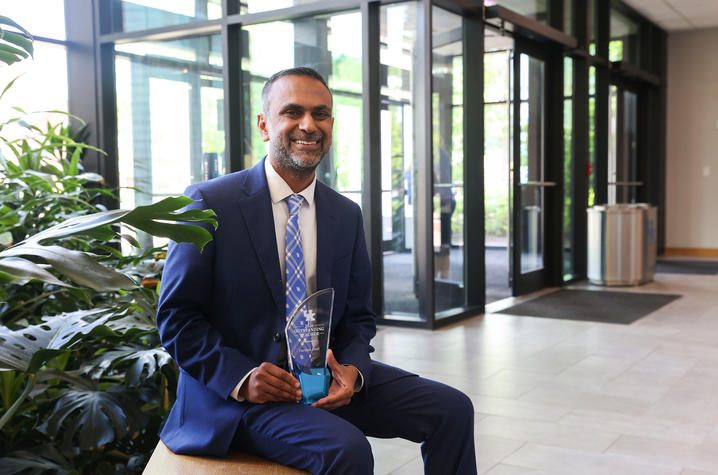 A person in a blue suit and University of Kentucky-patterned tie sits on a bench in a bright lobby, holding a glass award.