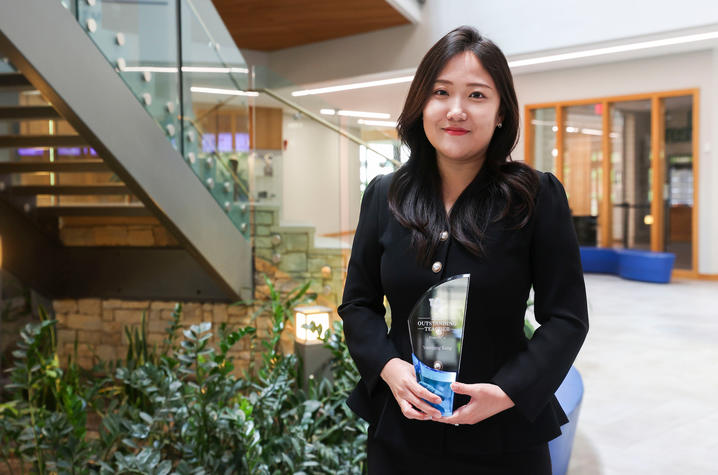Person in a black outfit standing in a modern building lobby and holding a clear glass award with a blue base. A staircase with glass railings, indoor plants, and seating areas are visible in the background. 
