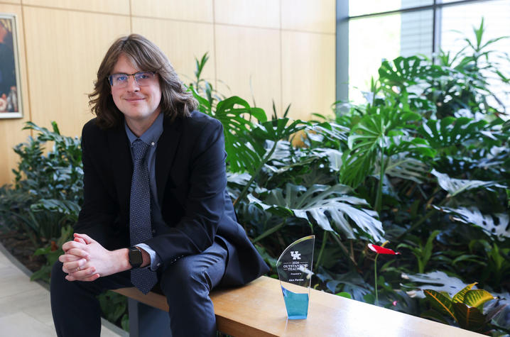 Person with shoulder-length hair wearing a dark jacket and glasses sits on a bench beside a small glass award, with large green plants behind them indoors.