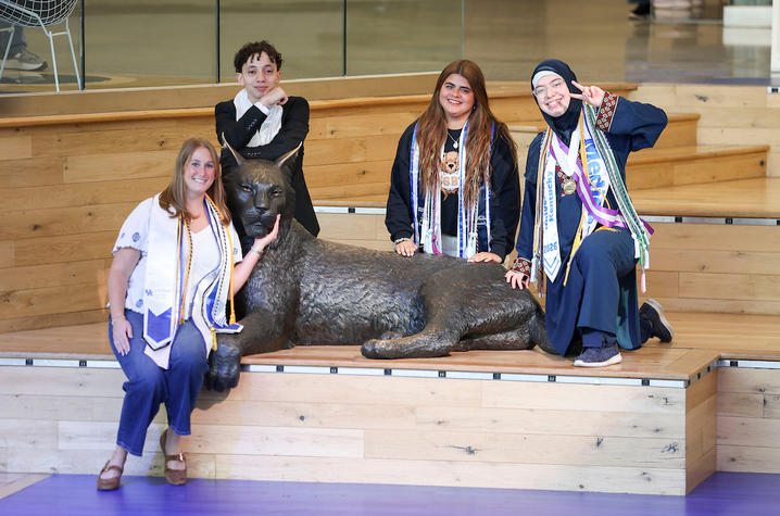 Four students pose with graduation stoles around the university’s Wildcat statue inside a campus building.