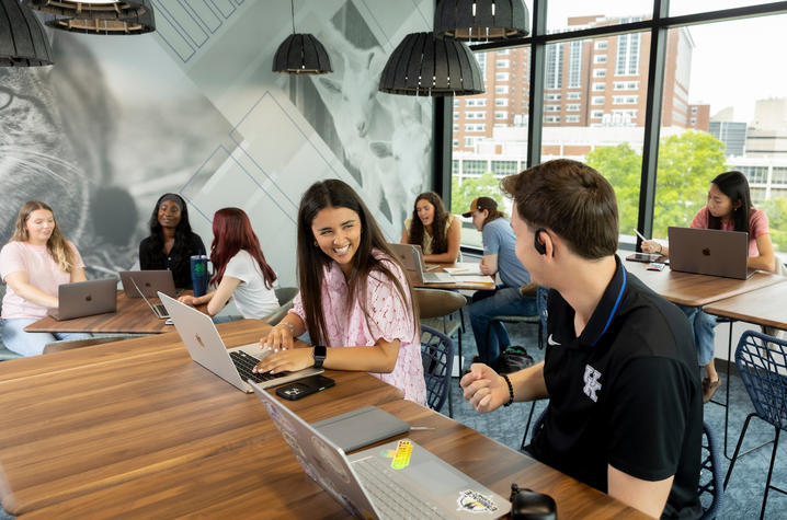 Students work and talk together around tables with laptops in a bright campus study space.