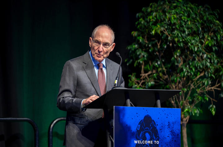 Adult in a gray suit and patterned tie speaks at a podium with a microphone, looking toward the audience. A plant and dark stage background frame the scene.