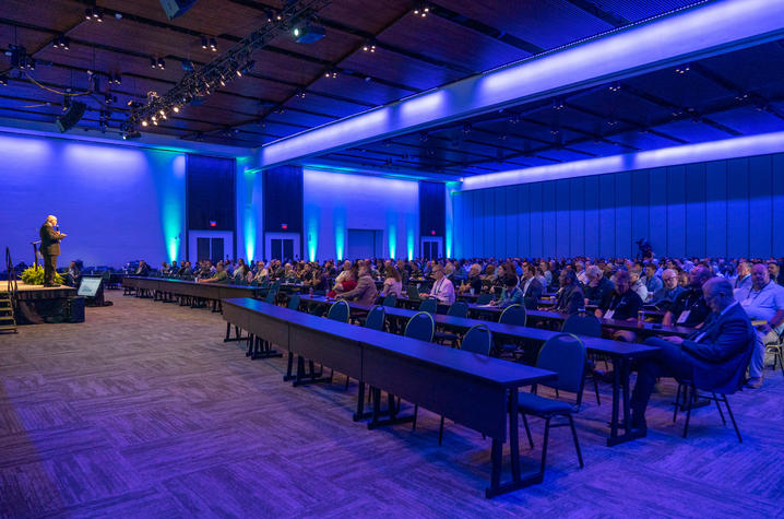Large conference room filled with seated attendees facing a speaker at the front. Blue and green lighting illuminates the space, with rows of tables and a stage presentation underway.