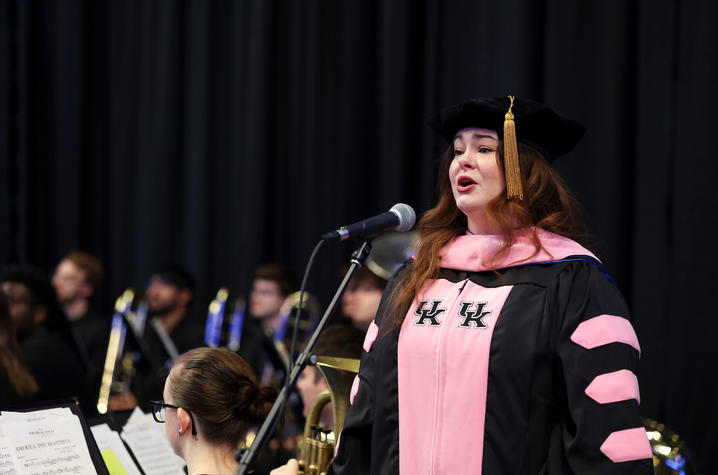A commencement speaker stands at a podium in academic regalia with musicians seated behind during a ceremony.
