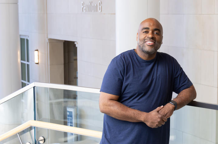 Photo of patient Tyrone Vinegar A person in a navy T-shirt leans on a glass railing inside a bright, modern building with light-colored walls and windows in the background.