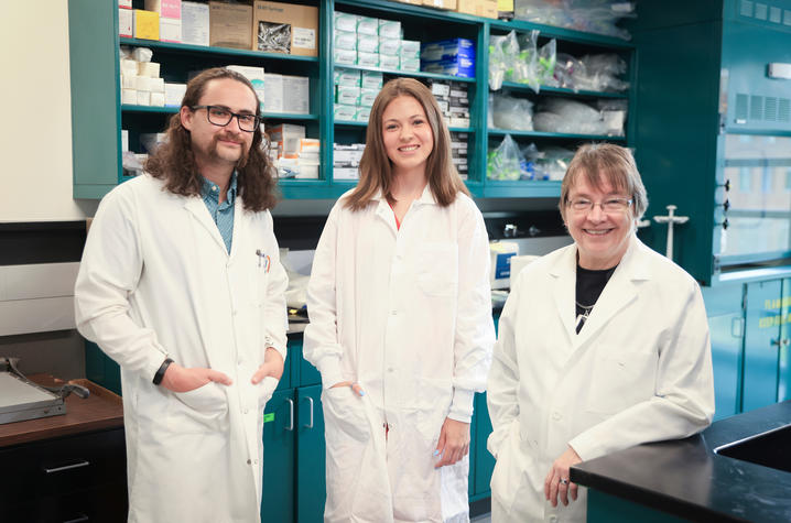 Three researchers wearing white lab coats stand together smiling in a laboratory filled with shelves of scientific supplies and equipment. The group poses in front of workstations and storage cabinets in a bright research setting.