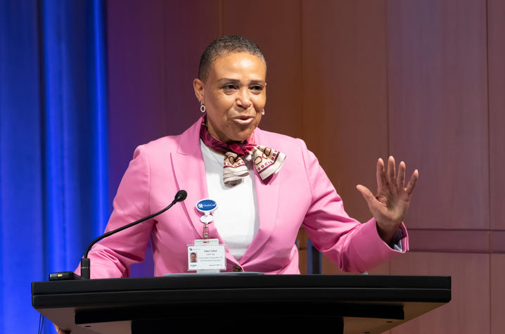 Photo of Tukea Talbert A person stands at a podium speaking into a microphone in a formal auditorium setting. The person wears a bright pink blazer with a patterned scarf tied at the neck and a name badge clipped to the lapel.