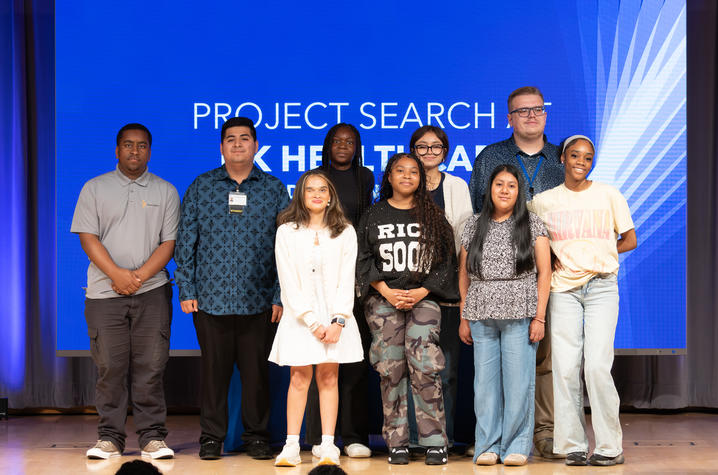 Photo of Project SEARCH graduates Seven young participants stand in a line on a stage in front of a large blue screen displaying the words “PROJECT SEARCH UK HealthCare.”