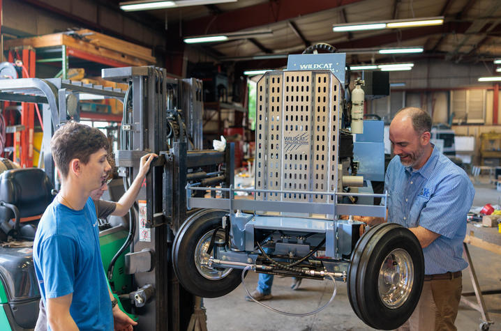 “Wildcat” tractor raised Two people work on a small, custom-built “Wildcat” tractor raised on a forklift inside a workshop. One person stands beside the forklift mast while another adjusts components near the front wheel assembly.