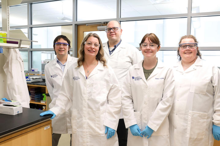 Five researchers wearing lab coats, safety glasses and gloves stand together in a University of Kentucky laboratory.