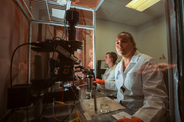 Two researchers wearing lab coats and protective gloves work with specialized laboratory equipment in a research setting. One is seated in the foreground operating an imaging system, while the second works at a computer station.