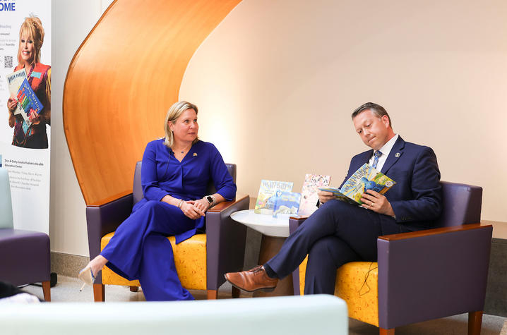 Two adults seated in a healthcare space read children’s books during an event, with additional books displayed nearby.