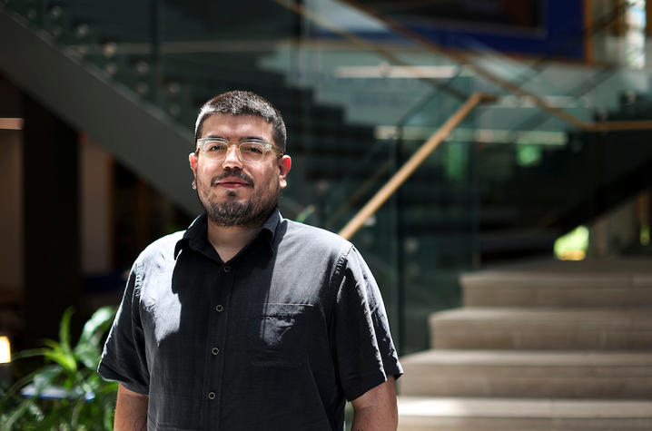Person in a black shirt stands outdoors near a modern building with glass and concrete steps in the background.