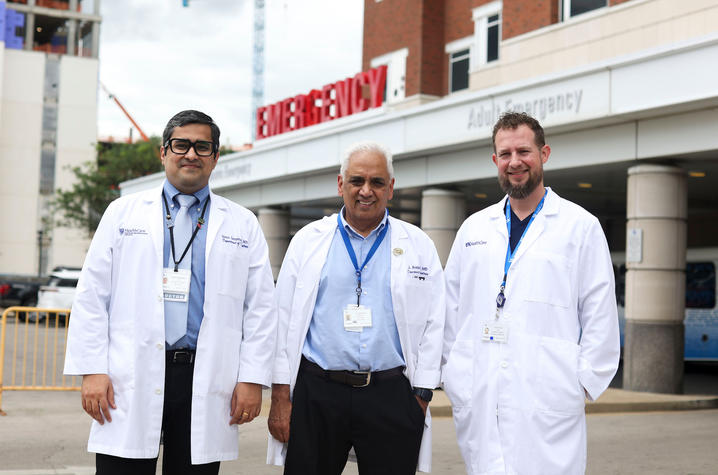 Three physicians wearing white lab coats stand outside the emergency department entrance at a hospital. The group poses together in front of the emergency care facility, highlighting a clinical and academic health care setting.