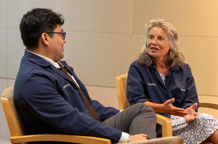 Two people participate in an interview at UK HealthCare. Two people seated in wooden chairs during an interview. Both are wearing blue UK HealthCare jackets and clip-on microphones. One person is speaking with hands raised while the other listens.