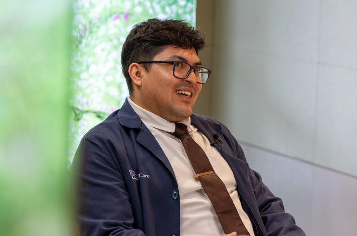 Clinical Pastoral Education participant during an interview at UK HealthCare. A person wearing glasses, a white shirt, tie and blue UK HealthCare jacket sits in a chair during an interview. A clip-on microphone is attached to the jacket, and a blurred green foreground element appears along the left side of the image.