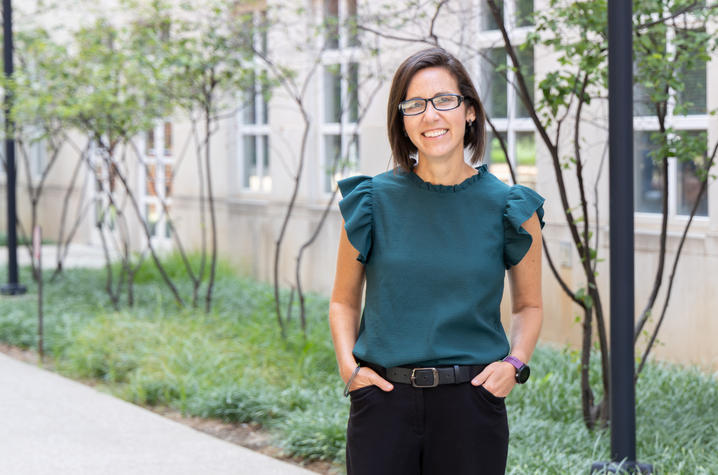 Photo of Christen Page in front of the College of Health Sciences A person stands on a landscaped campus walkway outside a building, wearing glasses and a dark green blouse, with trees and greenery in the background.