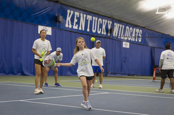 A young participant swings a tennis racquet to return a ball during a youth tennis clinic at the University of Kentucky indoor tennis facility. Coaches and volunteers in matching event T-shirts stand nearby on the court, with large “Kentucky Wildcats” sig