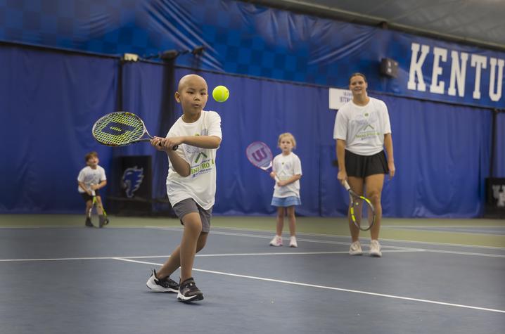 A child swings a tennis racquet to hit a bright yellow tennis ball on an indoor court at the University of Kentucky. Three other young tennis players and an instructor stand in the background holding racquets. Blue court curtains and large “KENTUCKY” lett