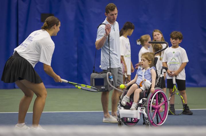 A young participant using a wheelchair holds a tennis ball while taking part in a tennis activity at the University of Kentucky indoor tennis facility. An instructor with a tennis racquet stands nearby, while other children and volunteers in Mini Pro Am T