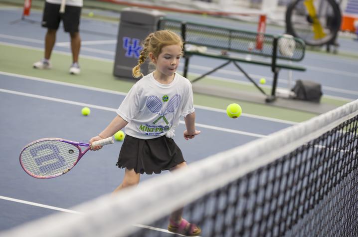 A young participant holds a tennis racquet while tracking a tennis ball near the net during a Mini Pro Am tennis activity at the University of Kentucky indoor tennis facility. Several tennis balls are scattered across the court in the background.