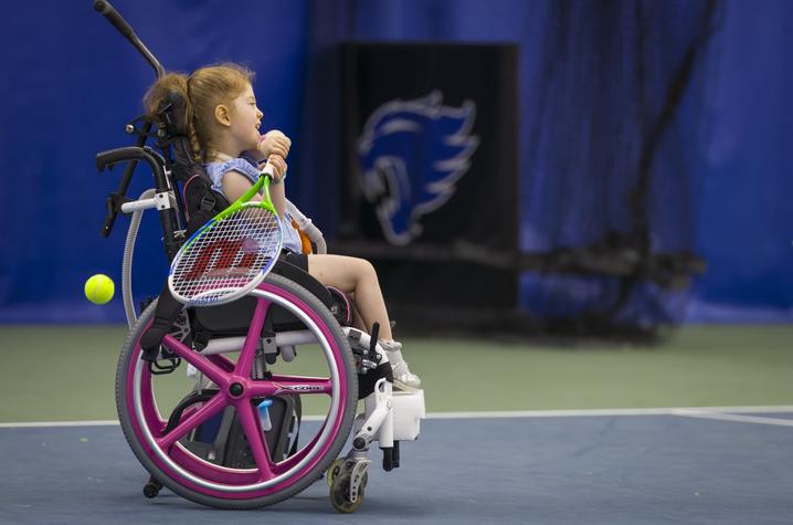 A young participant using a wheelchair holds a tennis racquet while taking part in a tennis activity on an indoor court at the University of Kentucky. A tennis ball is visible nearby, with blue UK-branded court backdrops in the background.