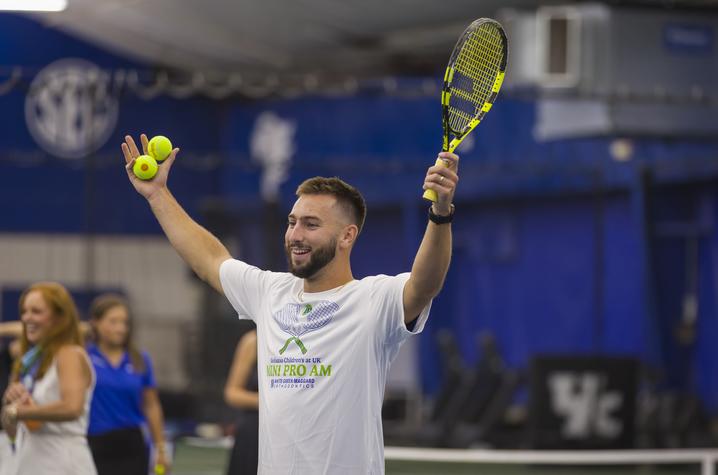 A volunteer holds up a tennis racquet and tennis balls while leading an activity during the Mini Pro Am event at the University of Kentucky indoor tennis facility. Other participants and spectators are visible in the background.