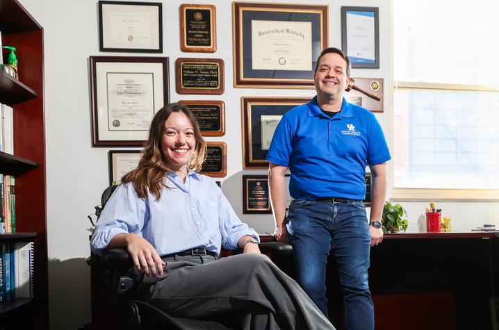 Researchers pose in an office displaying academic diplomas, awards and professional plaques on the wall. One person sits at a desk while another stands nearby, illustrating a collaborative research or clinical research team in a university setting.