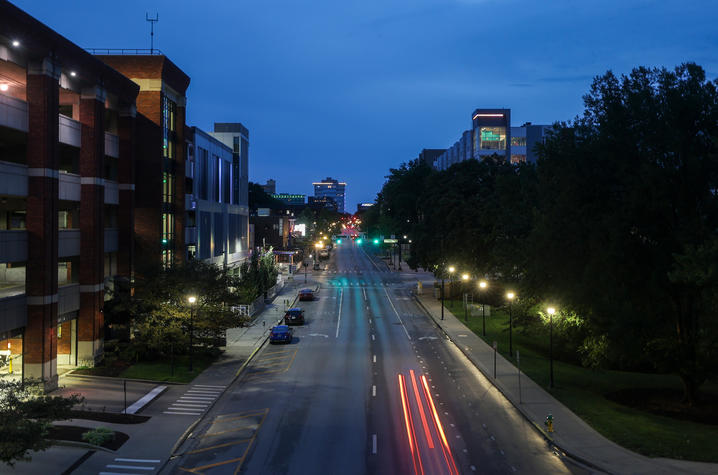 Elevated view of a campus street at dusk, with illuminated buildings and trees lining the road as a vehicle’s red taillights streak through the foreground.