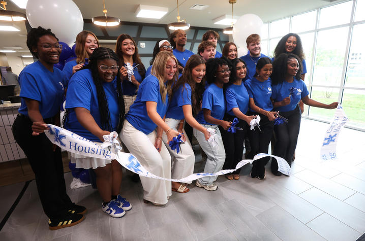 Group of students and university leaders in blue shirts gather indoors for a ribbon-cutting ceremony, holding ceremonial scissors and ribbon beneath white balloons.