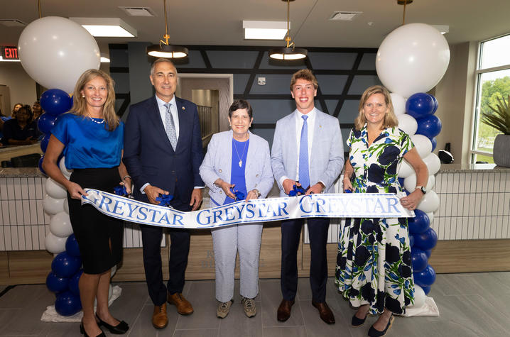 Four people stand behind a ceremonial ribbon inside a bright lobby during a ribbon-cutting event. Blue and white balloon columns flank the group, and the ribbon reads “GREYSTAR.” Tables and guests are visible in the background.