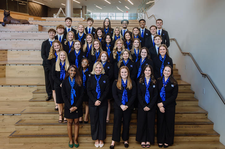 photo of 2026-27 Alumni Ambassadors A large group of students wearing matching professional attire and blue scarves pose on tiered wooden steps inside a campus building.